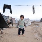 Criança caminha descalça entre tendas improvisadas e roupas estendidas em um campo de refugiados.