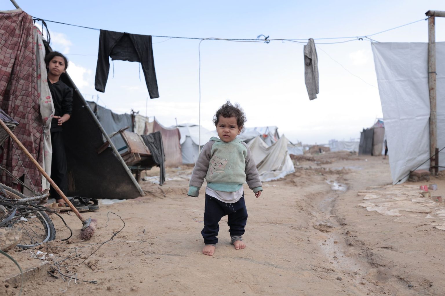 Criança caminha descalça entre tendas improvisadas e roupas estendidas em um campo de refugiados.