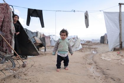 Criança caminha descalça entre tendas improvisadas e roupas estendidas em um campo de refugiados.
