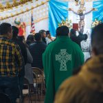 Sacerdote conduz celebração religiosa em uma capela, enquanto fiéis rezam em pé.
