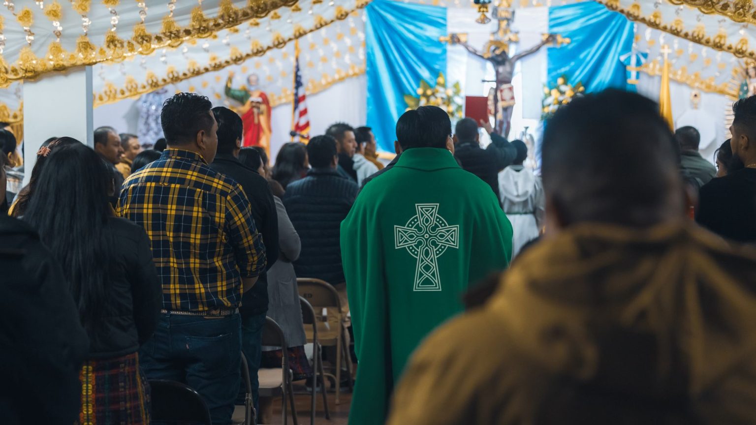 Sacerdote conduz celebração religiosa em uma capela, enquanto fiéis rezam em pé.