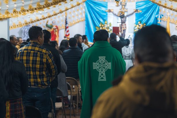 Sacerdote conduz celebração religiosa em uma capela, enquanto fiéis rezam em pé.