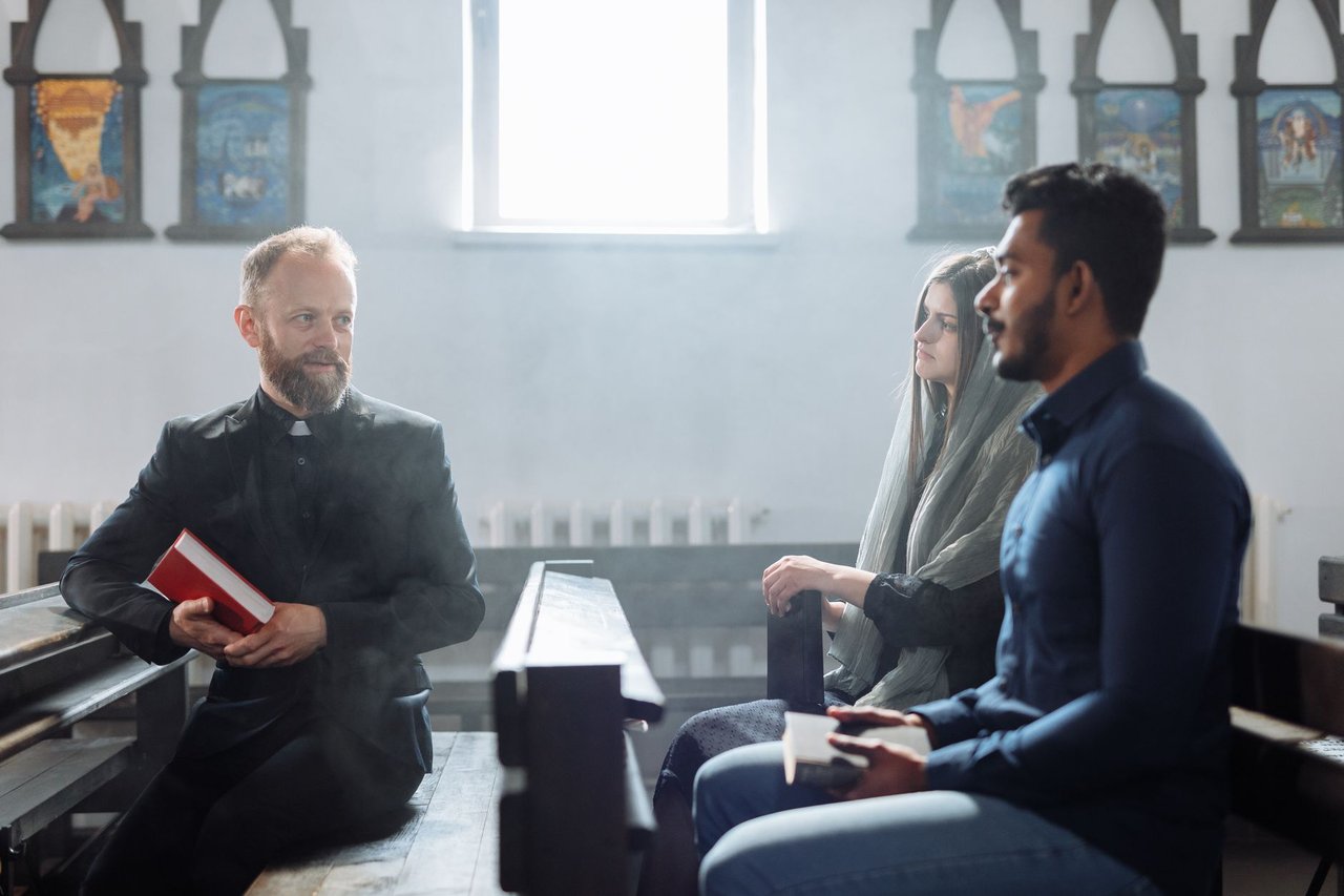Padre conversa com duas pessoas no interior de uma igreja, em uma cena de diálogo e convivência comunitária.